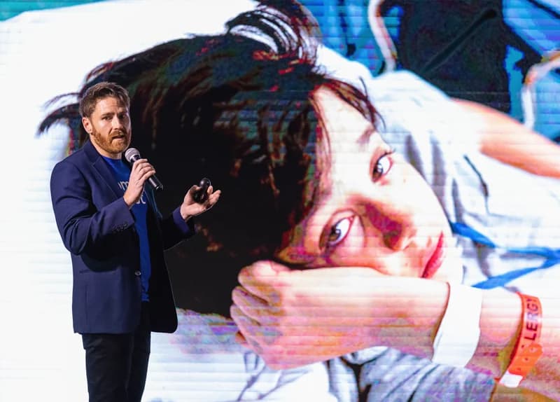 Taylor Hobbs on stage, telling the Comfort Quest story in front of a photo of a child in hospital